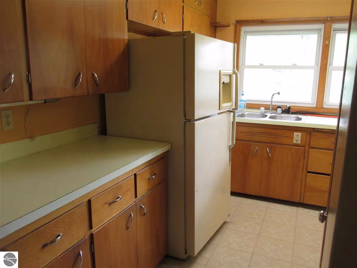 A small kitchen with wooden cabinets, a beige refrigerator, a double sink under a window, and a beige countertop.