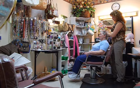 A woman is sitting in a salon chair while another woman styles her hair in a small room filled with various accessories such as handbags, necklaces, and scarves displayed on shelves and hanging on the wall.