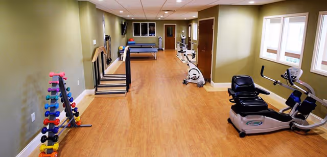A fitness room with wooden flooring and light green walls featuring exercise equipment including a recumbent bike, an upright bike, a set of colorful dumbbells on a rack, and a ping pong table at the far end. There are windows on the right wall and a door at the back of the room.