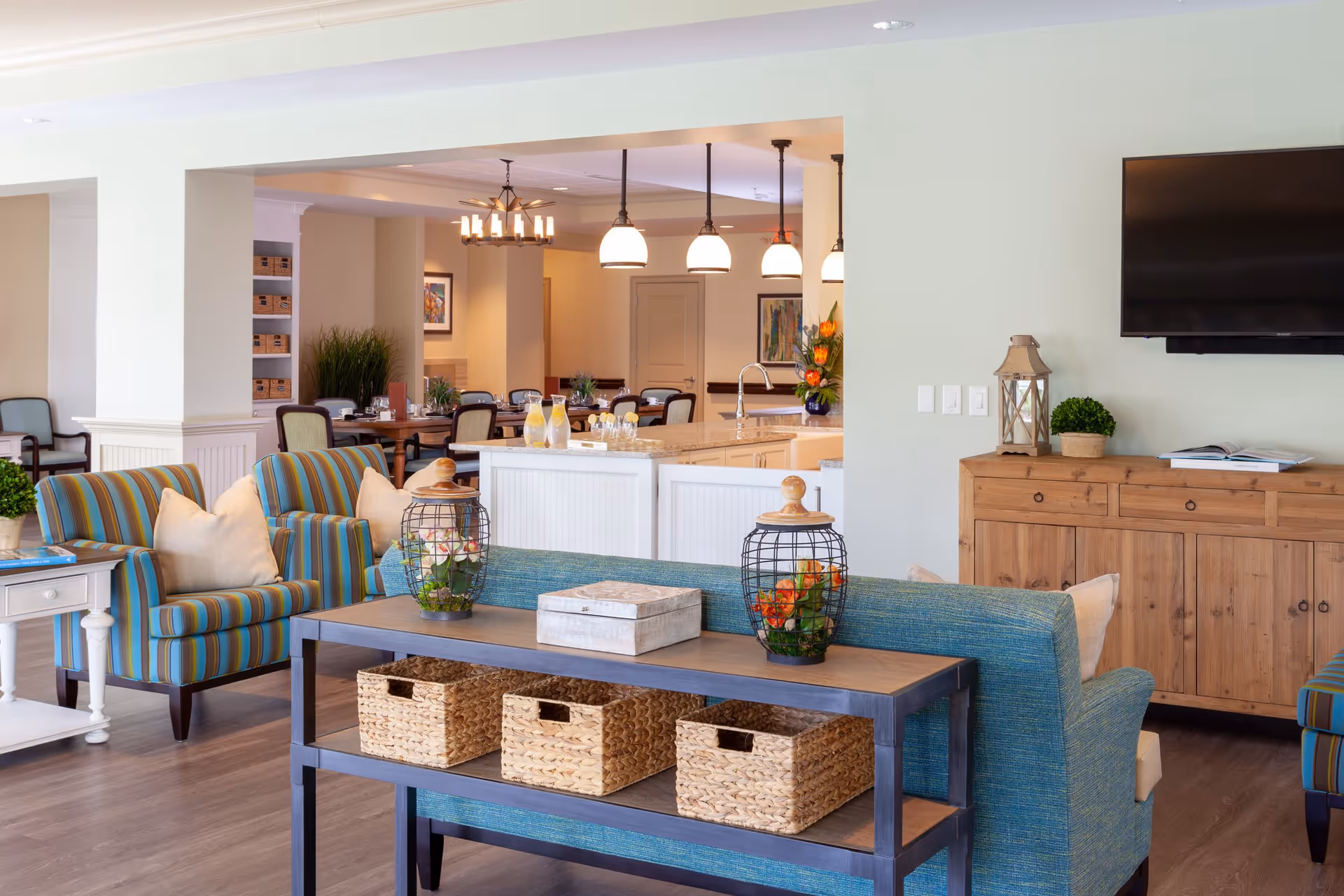 A bright and spacious living room area with blue and striped armchairs, a wooden console table with three woven baskets underneath, decorative lanterns with flowers, and a wooden cabinet with a flat-screen TV mounted above. In the background, there is a dining area with tables and chairs, pendant lights, and a kitchen island with a sink and pitchers of lemonade.
