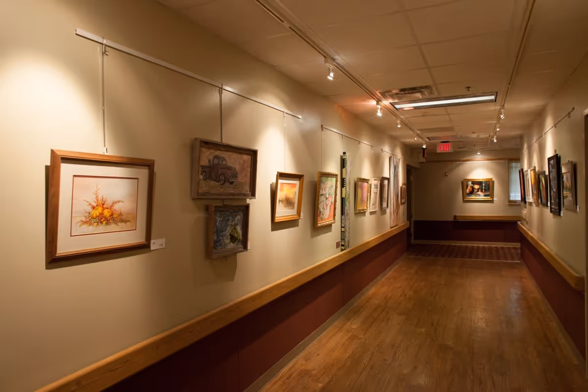 A well-lit hallway with wooden flooring and beige walls, featuring various framed paintings and artwork displayed on both sides. The hallway has a wooden handrail running along the walls and ceiling lights illuminating the artwork. An exit sign is visible at the far end.