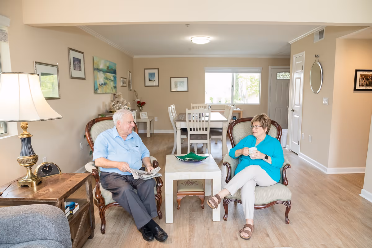 Two seniors sit in armchairs facing each other in a bright living room with a coffee table and a dining area visible in the background.