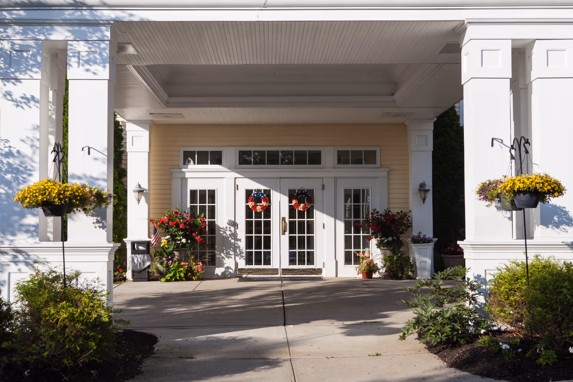 Entrance of Van Dyk Park Place facility with white double doors adorned with wreaths, flanked by large white columns and hanging flower baskets with yellow flowers. There are various potted plants and flowers near the doorway, and the area is well-lit with natural sunlight.