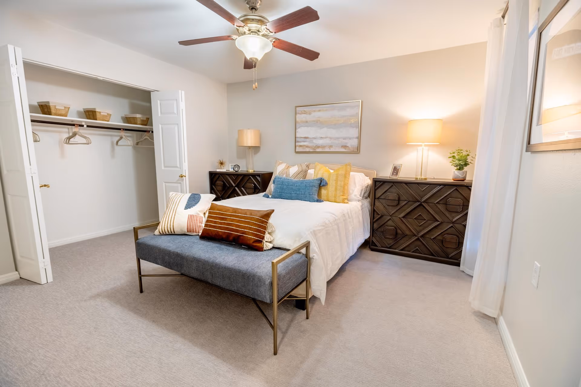 Well-lit bedroom with a bed topped with colorful pillows, a bench at the foot, an open closet, ceiling fan, and matching nightstands.