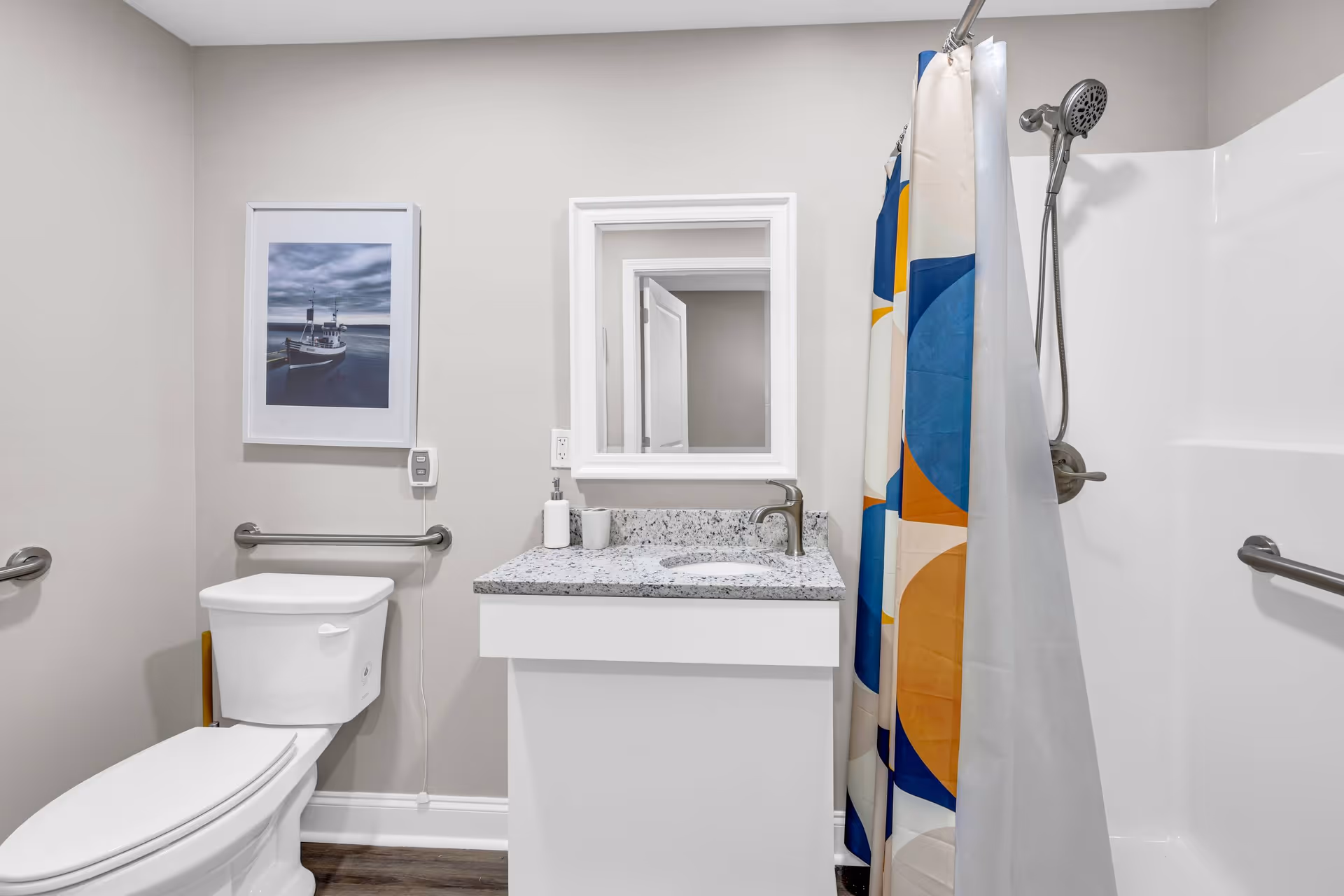 A clean and modern bathroom with a white toilet, a white vanity with a granite countertop and a sink, a mirror above the sink, a colorful shower curtain with blue, orange, and beige patterns, a showerhead, and grab bars on the walls. There is also a framed picture of a boat on the wall.