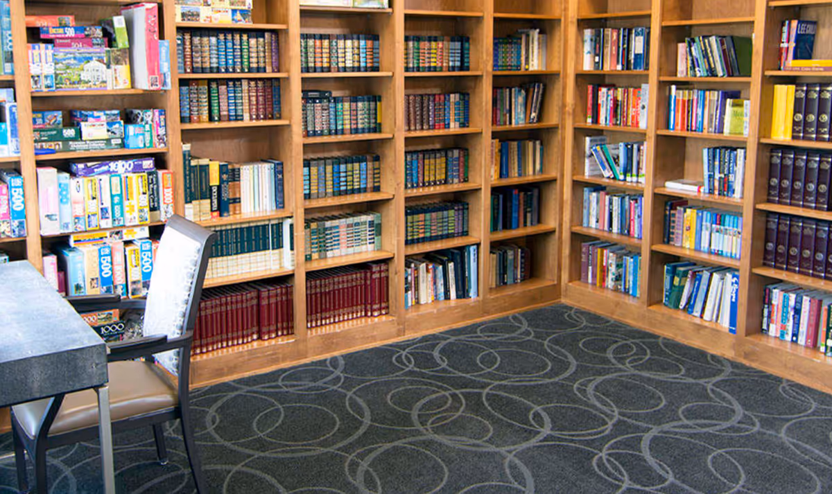 Corner of an indoor library or reading room with wooden bookshelves filled with books, a table and chair, and patterned carpet.
