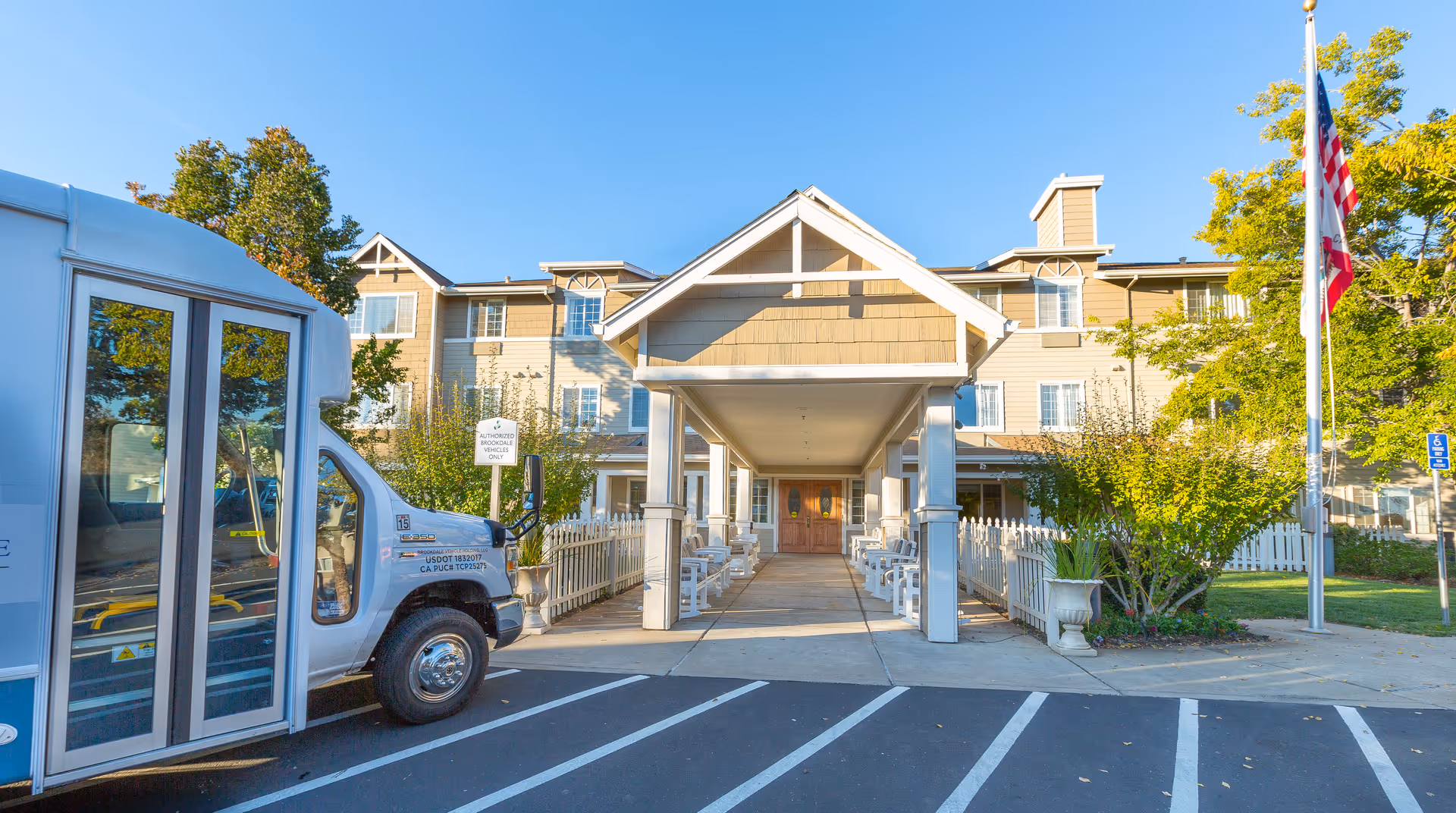 Exterior view of Brookdale Auburn senior living facility entrance with a covered walkway, white picket fences, benches, and a parked shuttle bus on the left. There is an American flag on the right side and trees surrounding the building under a clear blue sky.