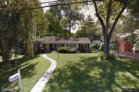 Single-story brick house with a front porch, surrounded by trees and a well-maintained lawn with a curved concrete walkway leading to the front door. There is a white mailbox near the sidewalk.