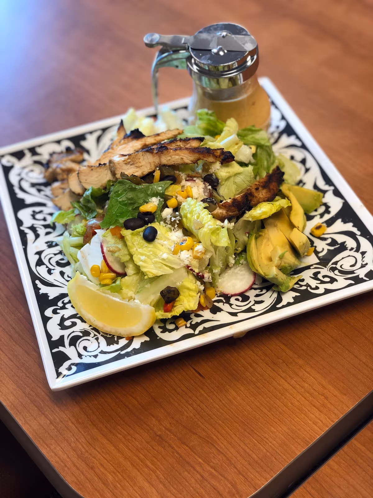 A plate of salad with grilled chicken strips, lettuce, corn, black beans, radish slices, avocado slices, and a lemon wedge, served on a decorative black and white square plate with a small glass container of dressing on the side, placed on a wooden table.