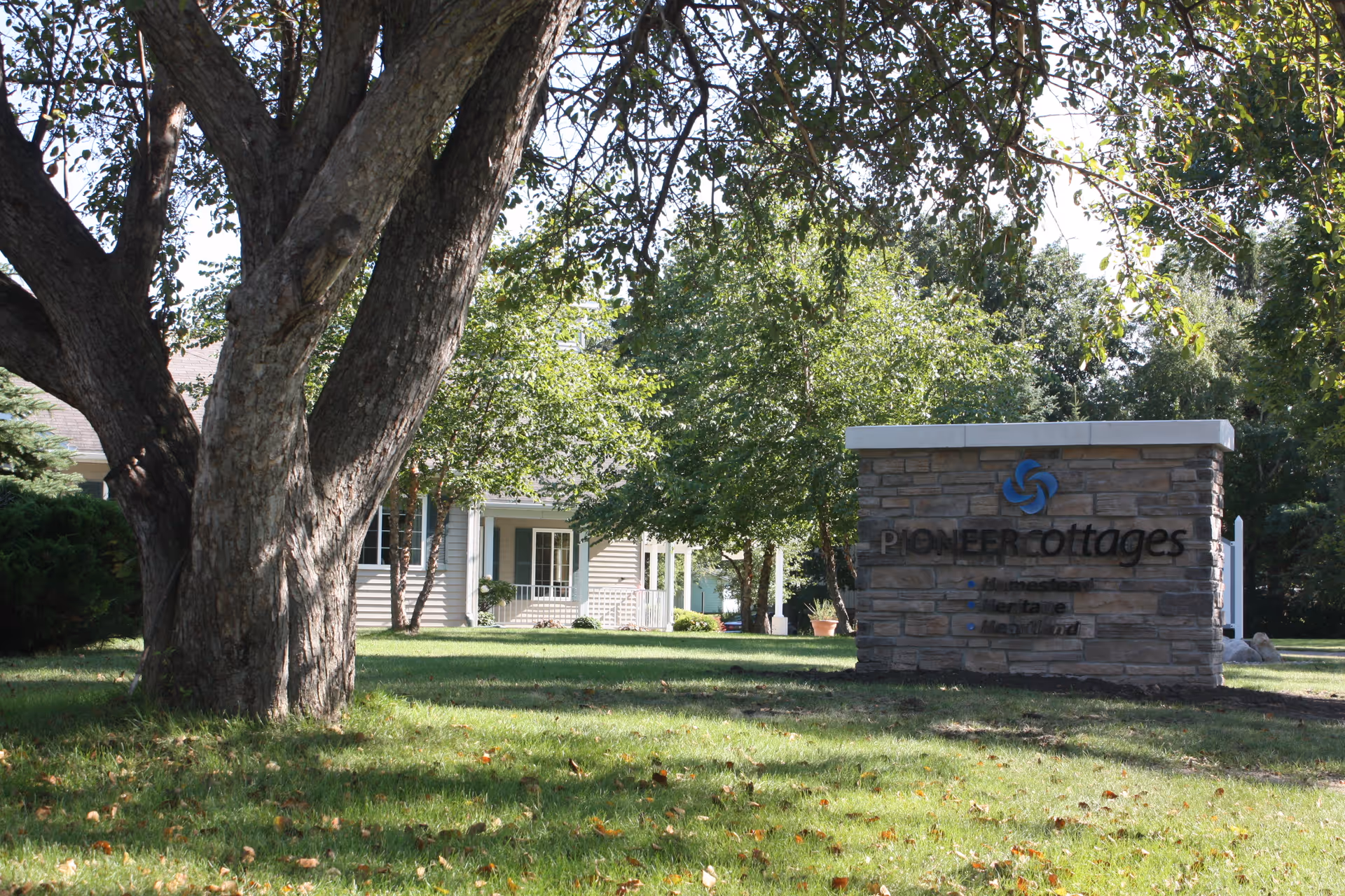 A large tree in the foreground with a grassy lawn and several smaller trees. In the background, there is a light-colored building with a porch. To the right, there is a stone sign that reads 'Pioneer Cottages Memory Care' with a blue logo above the text.