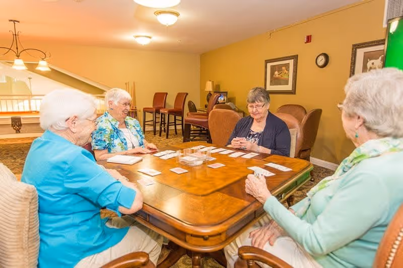 Four elderly women sitting around a wooden table playing cards in a well-lit room with beige walls and carpeted floor. The room has chairs, framed pictures on the wall, and a clock. The atmosphere appears warm and social.
