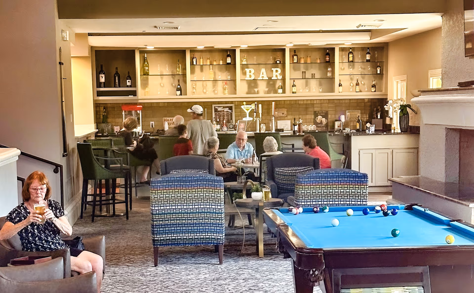 A senior living facility common area featuring a pool table in the foreground, several armchairs and small tables, and a bar with shelves of bottles and glasses in the background. Several elderly people are seated and standing around the bar and seating area, engaging in conversation and relaxing.