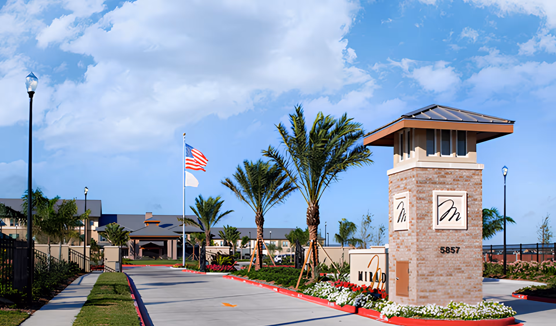 Driveway entrance and landscaped front sign with a brick tower, palm trees, and an American flag at a retirement community.