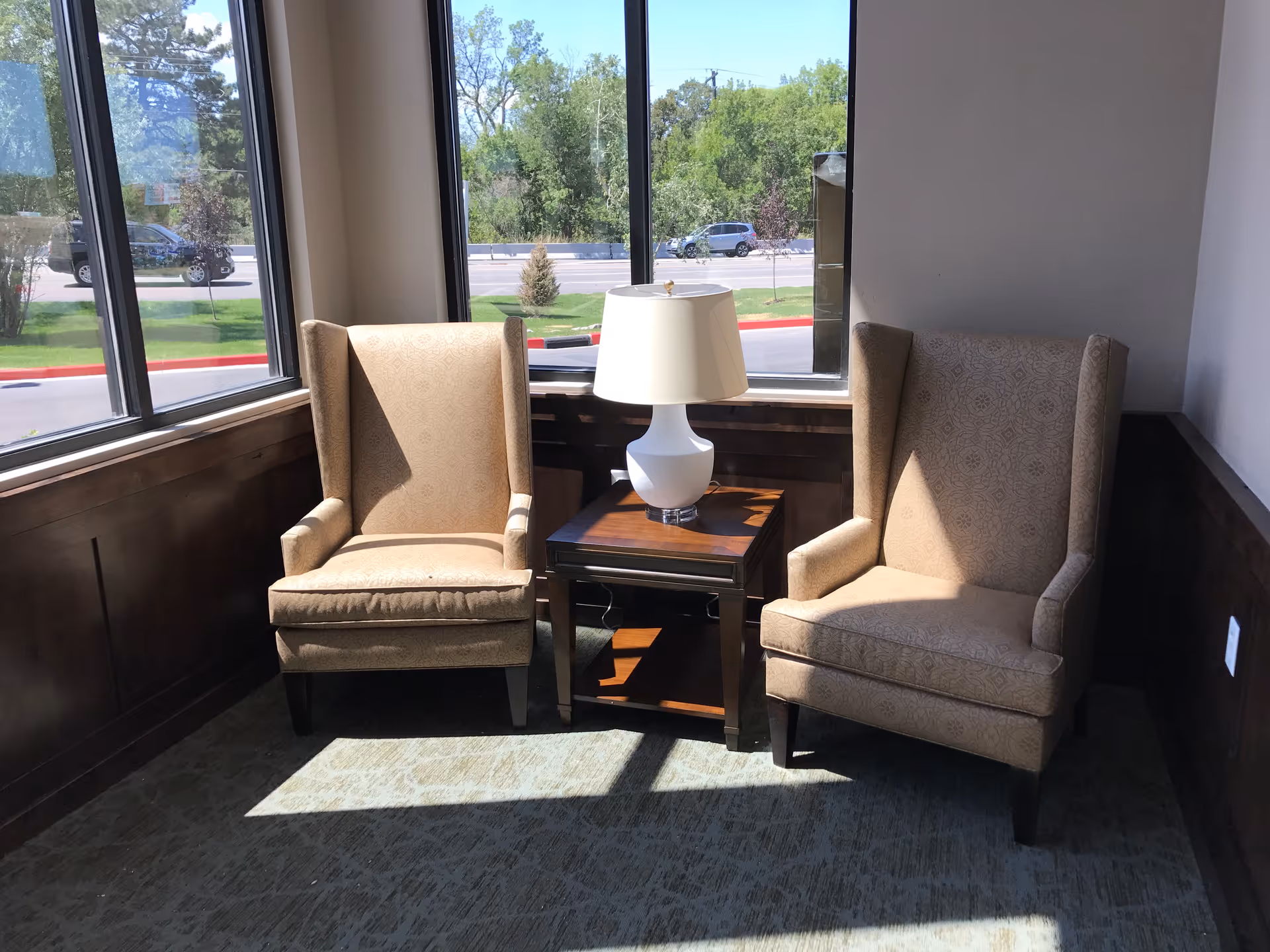 Two beige upholstered armchairs with patterned fabric are positioned on either side of a small wooden side table with a white table lamp. The seating area is next to large windows that let in natural light, showing a view of trees, grass, and parked cars outside.
