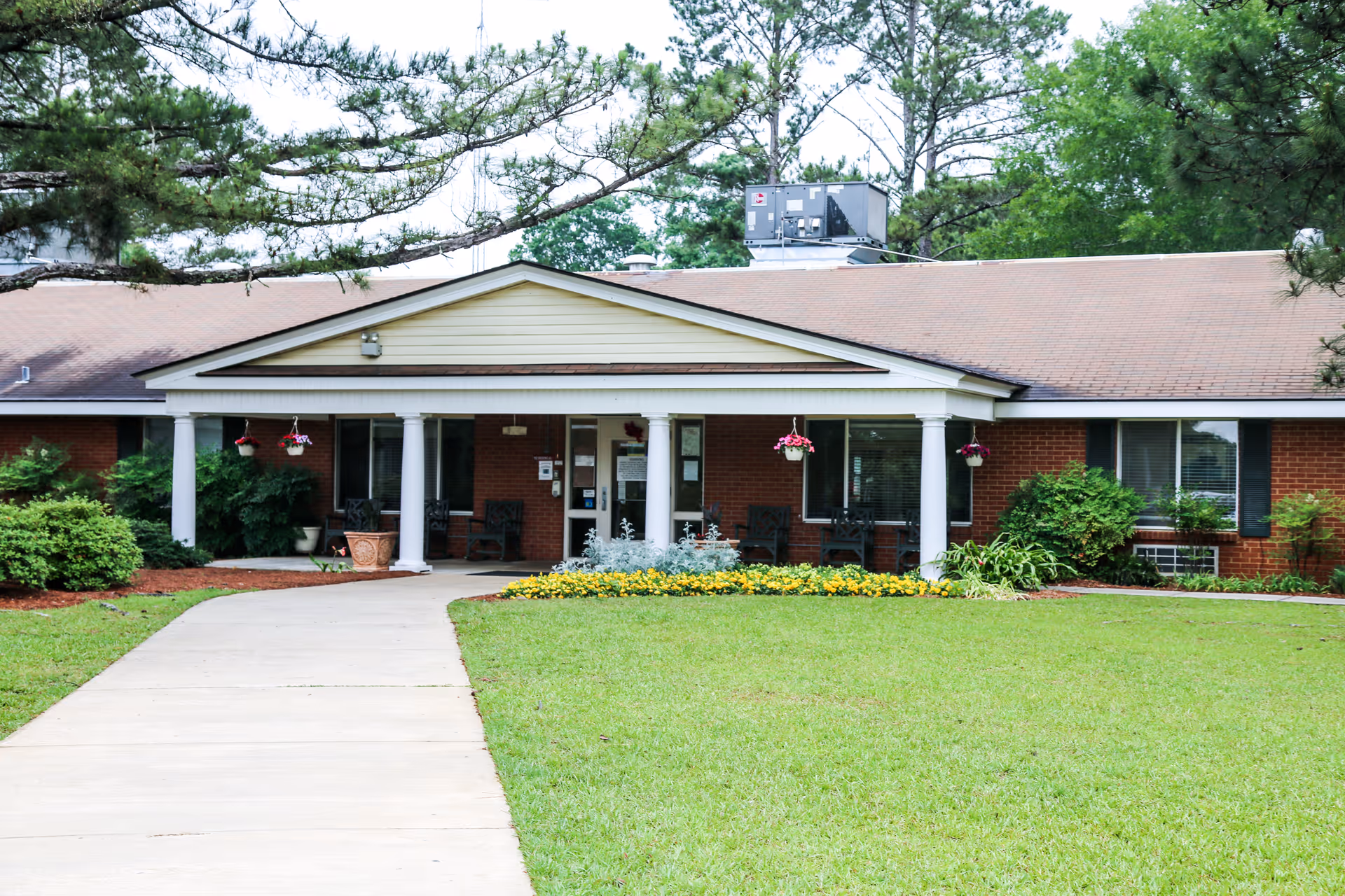 Front exterior view of a single-story brick building with a covered entrance supported by white columns. There are hanging flower pots and chairs on the porch. A concrete walkway leads up to the entrance, surrounded by a well-maintained lawn and landscaping with bushes and flowers. Tall trees are visible in the background.
