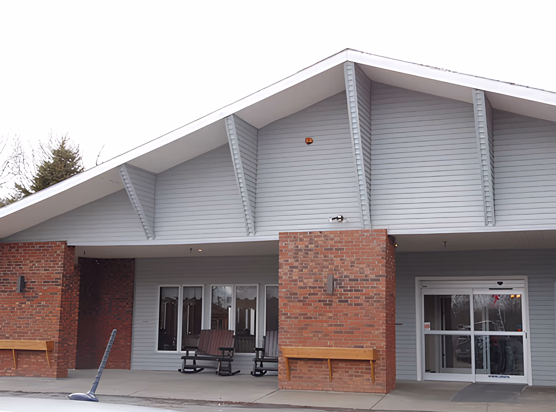 Covered front entrance of a single-story building with brick columns, rocking chairs on a porch and sliding glass doors.