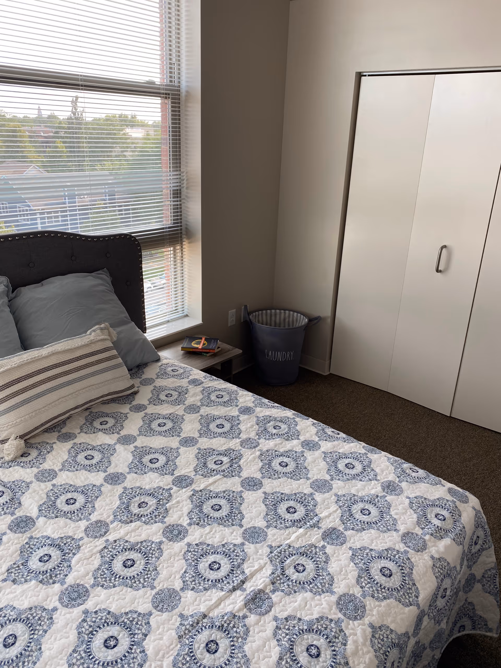 A bedroom with a bed covered in a white and blue patterned quilt, a striped pillow, and two gray pillows. Next to the bed is a small wooden side table with a book on it. There is a large window with blinds partially open, showing a view of trees and houses outside. In the corner of the room is a gray laundry basket labeled 'LAUNDRY'. A white closet with sliding doors is visible on the right side of the image.