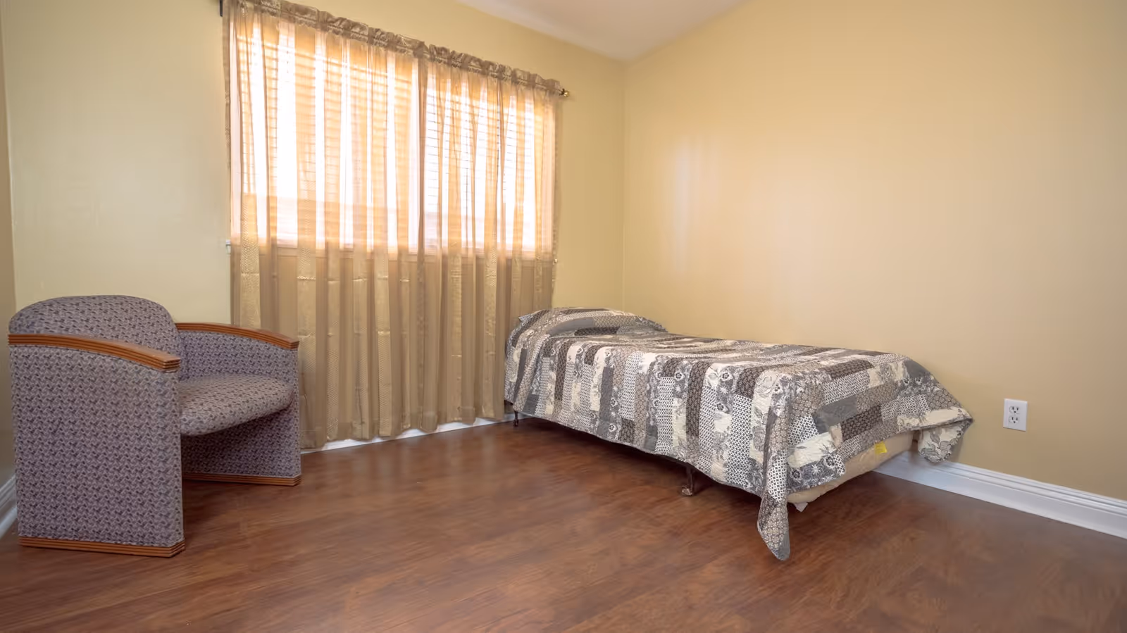 Small bedroom with a single bed covered by a patchwork quilt, an upholstered armchair, sheer window curtains, and wood flooring.
