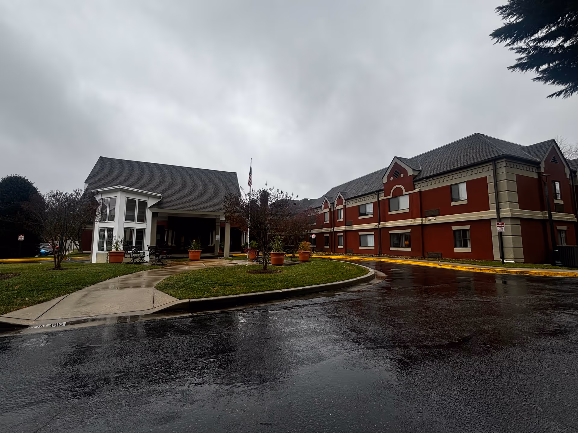 Exterior view of Aspenwood Senior Living Community on a cloudy day, showing a two-story red brick building with beige trim and a covered entrance with seating and potted plants. The driveway and sidewalk are wet from recent rain.