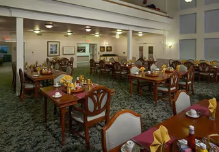 A spacious dining room with multiple wooden tables and chairs arranged neatly. Each table is set with folded yellow napkins, cups, saucers, and condiments. The room has a high ceiling with recessed lighting and framed artwork on the walls.