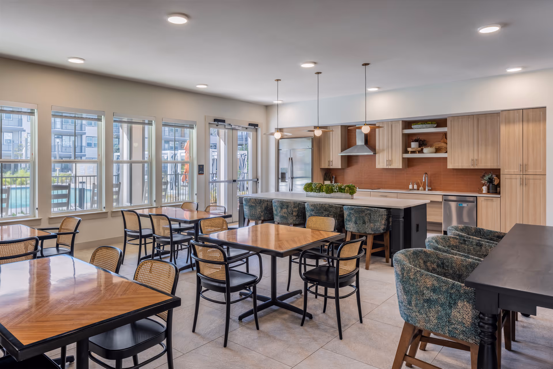 A bright and modern dining area with multiple wooden tables and black chairs with woven backs. The room features large windows letting in natural light and a view of an outdoor pool area. At the back, there is a kitchen with light wood cabinets, a stainless steel refrigerator, dishwasher, and a kitchen island with green cushioned bar stools. Pendant lights hang above the island.