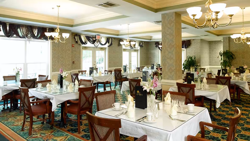 A well-lit dining room with multiple tables covered in white tablecloths, each set with glassware, cups, napkins, and floral centerpieces. The room features patterned carpet, chandeliers, large windows with draped curtains, and decorative wallpaper.