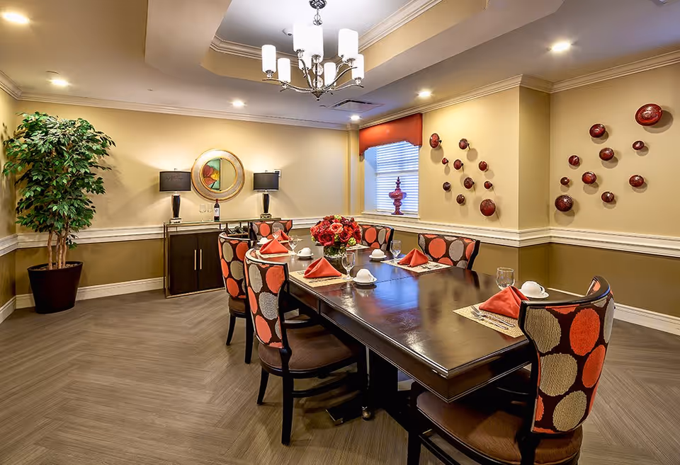 A well-lit dining room with a dark wooden table set for six people. The chairs have a colorful circular pattern in red, orange, and beige. The table is decorated with red napkins, white cups, glasses, and a centerpiece of red flowers. The room features beige walls with white trim, a large potted plant in the corner, a sideboard with two black lamps and a round mirror above it, and a window with a red valance. Red decorative wall art is displayed on one wall.