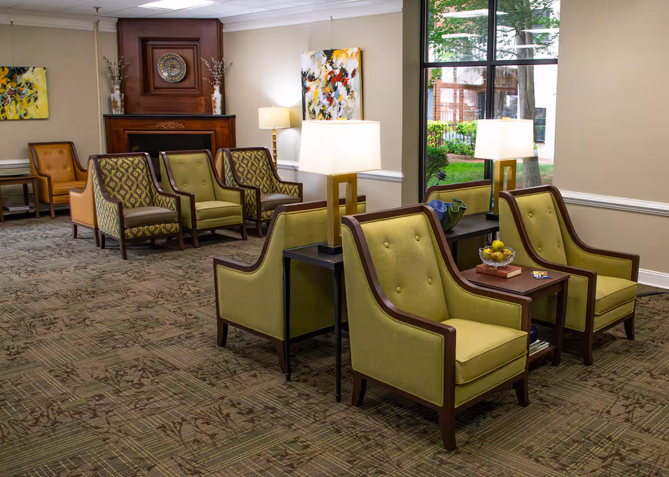 Bright senior living lounge with green upholstered armchairs arranged around side tables and lamps near a large window and fireplace.