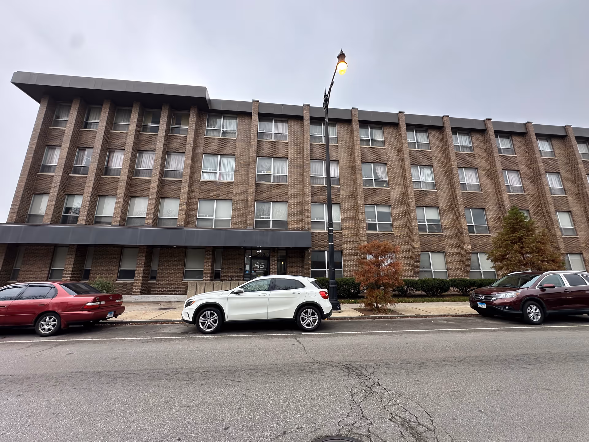 Front exterior view of a multi-story brick building with multiple windows, a black awning over the entrance, and three cars parked along the street in front. A streetlamp is lit near the entrance, and small trees and bushes line the sidewalk.