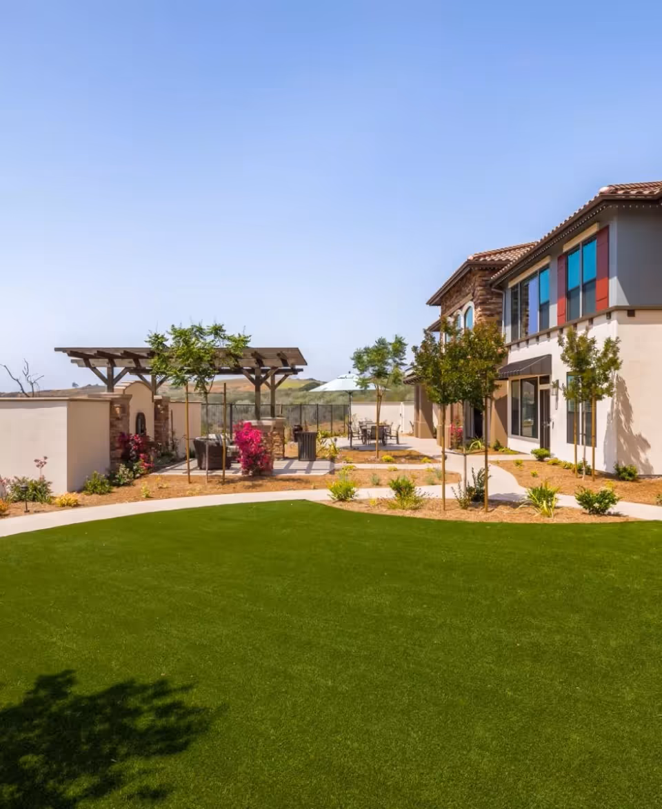 Courtyard with a green lawn, pergola seating area, landscaping and a two-story building with colorful windows under a clear blue sky.