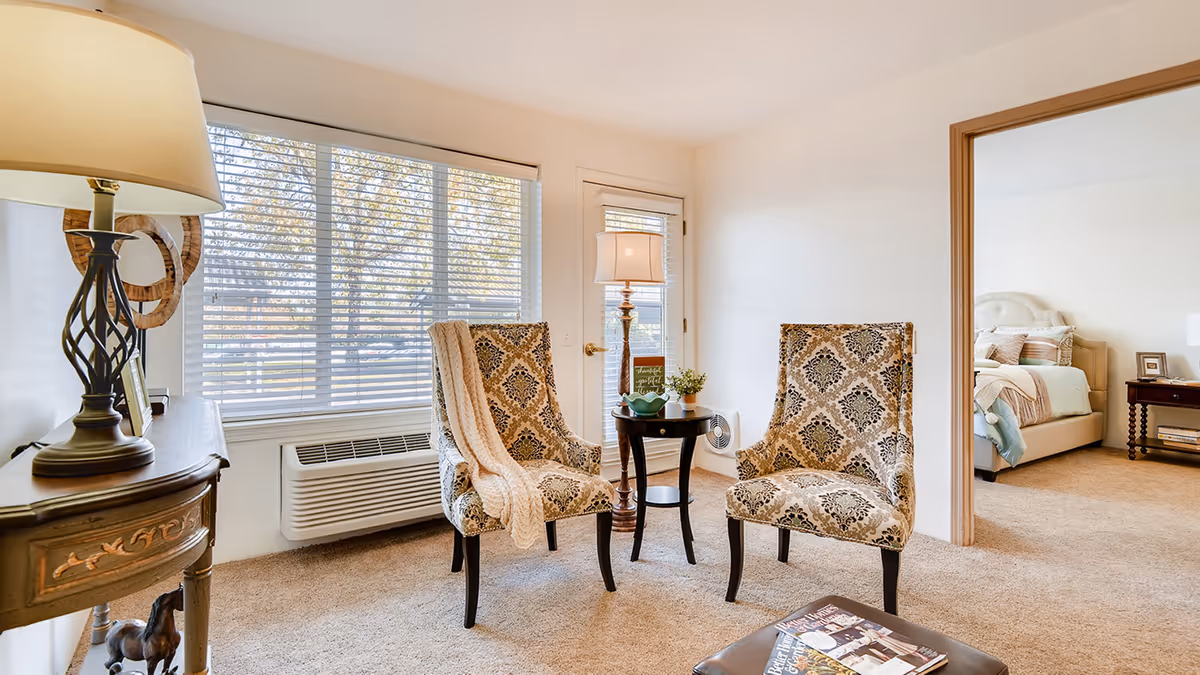 A cozy sitting area with two patterned armchairs and a small round table with a lamp and decorative items. A window with blinds lets in natural light, and a door is visible behind the chairs. To the right, an open doorway leads to a bedroom with a bed, nightstand, and lamp.