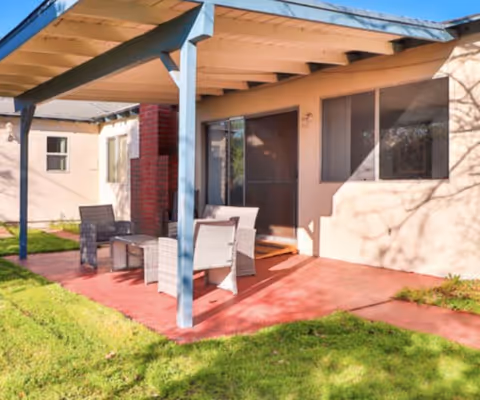 Outdoor patio area with a covered pergola attached to a building. The patio has a red concrete floor and is furnished with a small table and four wicker chairs. The surrounding area has green grass and the building has beige walls with windows and a sliding glass door.