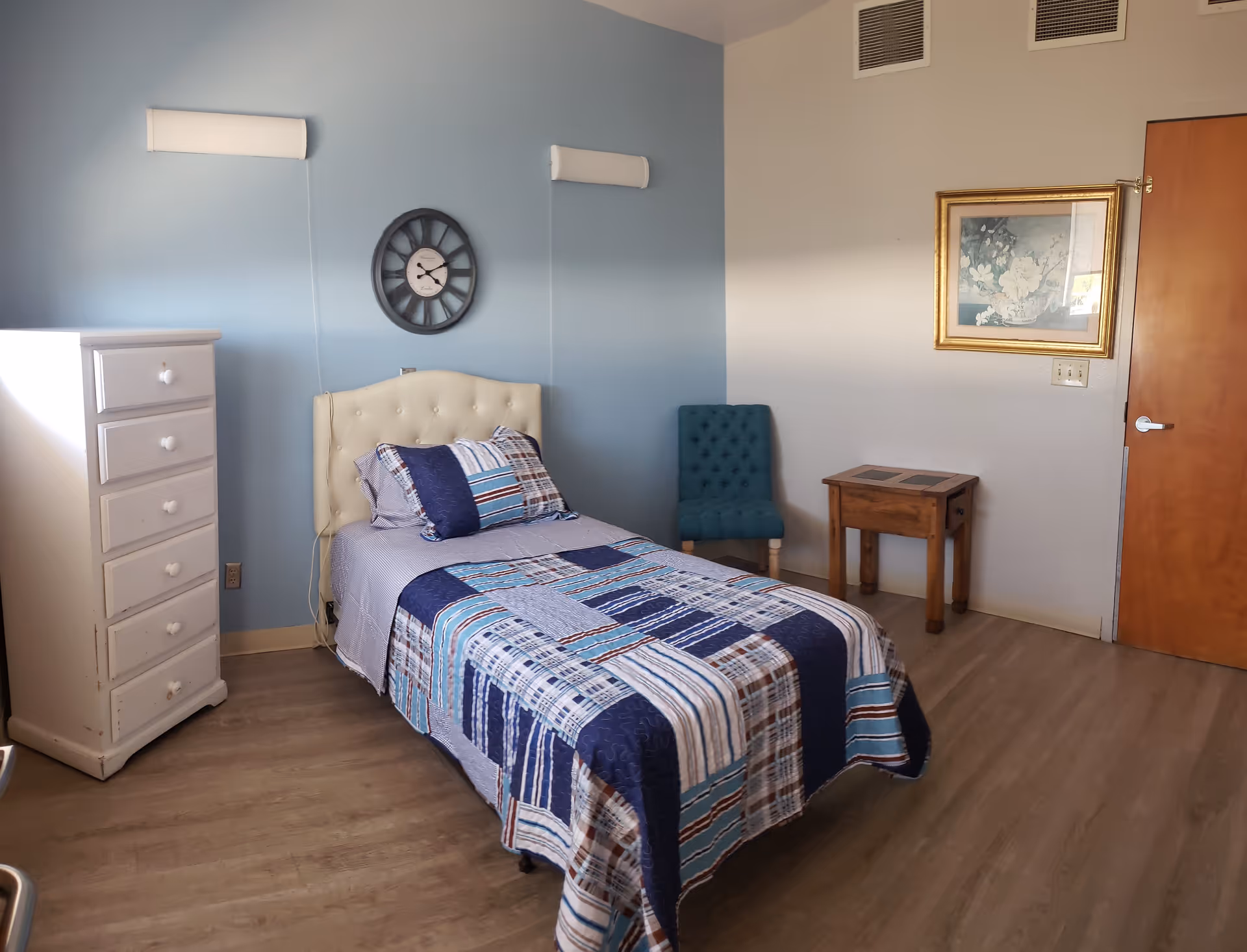 A neatly arranged bedroom with a single bed covered in a blue, white, and brown patchwork quilt. The bed has a tufted cream-colored headboard and is positioned against a light blue wall with a round black clock hanging above it. To the left of the bed is a white chest of drawers, and to the right is a small wooden side table and a teal upholstered chair. A framed floral picture hangs on the beige wall next to a wooden door. The floor is light wood laminate.