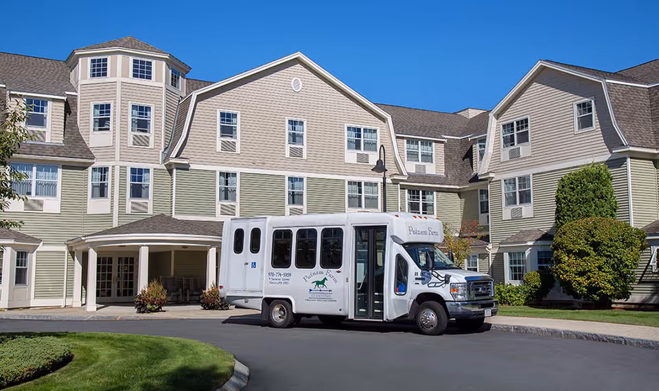 Exterior view of a multi-story senior living facility building with beige and green siding, multiple windows, and a covered entrance. A white shuttle bus with the text 'Putnam Farm' is parked in front of the building on a curved driveway. The sky is clear and blue, and there is well-maintained landscaping with grass and bushes.