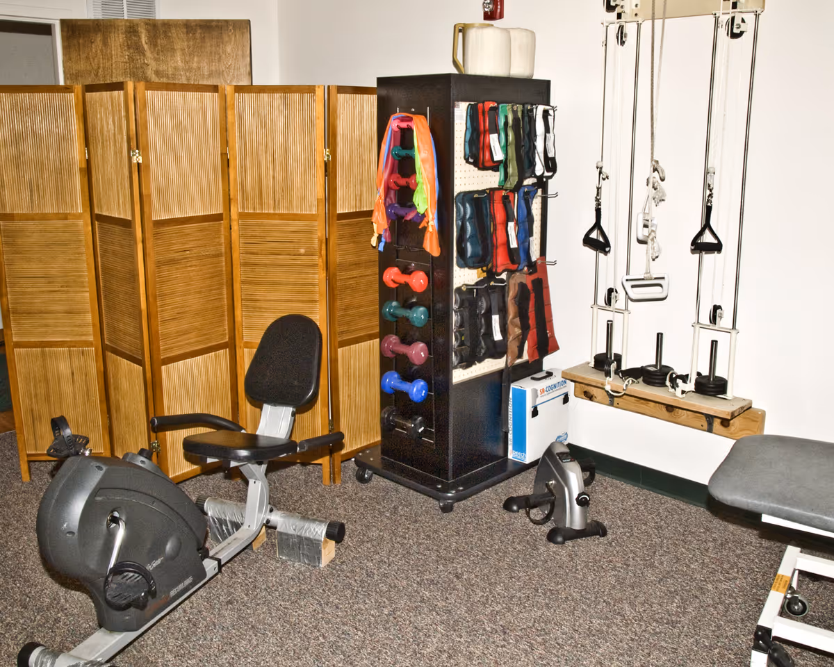 Small therapy/exercise room with a recumbent bike, dumbbell rack, resistance bands, pulley exercise system and a folding room divider.