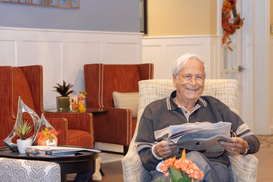An elderly man with white hair sitting comfortably in a beige armchair, smiling while holding a newspaper. Behind him are two red armchairs with a small table between them, decorated with a small plant and a pumpkin ornament. The room has light-colored walls with white wainscoting and a door with a fall-themed wreath.
