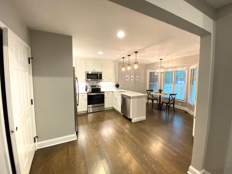 A modern kitchen and dining area with white cabinets, stainless steel appliances including a refrigerator, stove, microwave, and dishwasher. The kitchen has a white island with pendant lights hanging above it. The dining area features a round table with four chairs and large windows with blinds, allowing natural light to enter. The floor is wooden and the walls are painted light gray.