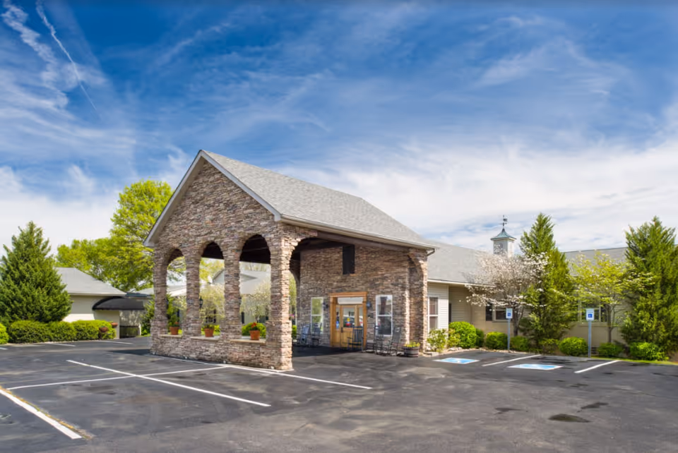Front entrance of a single-story senior living building with a stone portico, parking lot and landscaped trees under a blue sky.