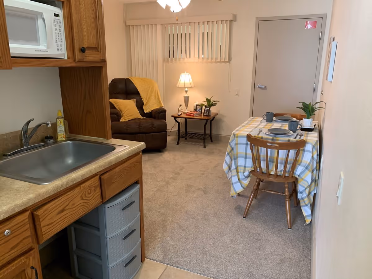 A cozy assisted living apartment interior featuring a small kitchen area with a sink and microwave on the left, a dining table with two chairs covered with a checkered tablecloth on the right, and a comfortable recliner chair with a yellow throw blanket and pillow in the background next to a side table with a lamp and decorative plants.