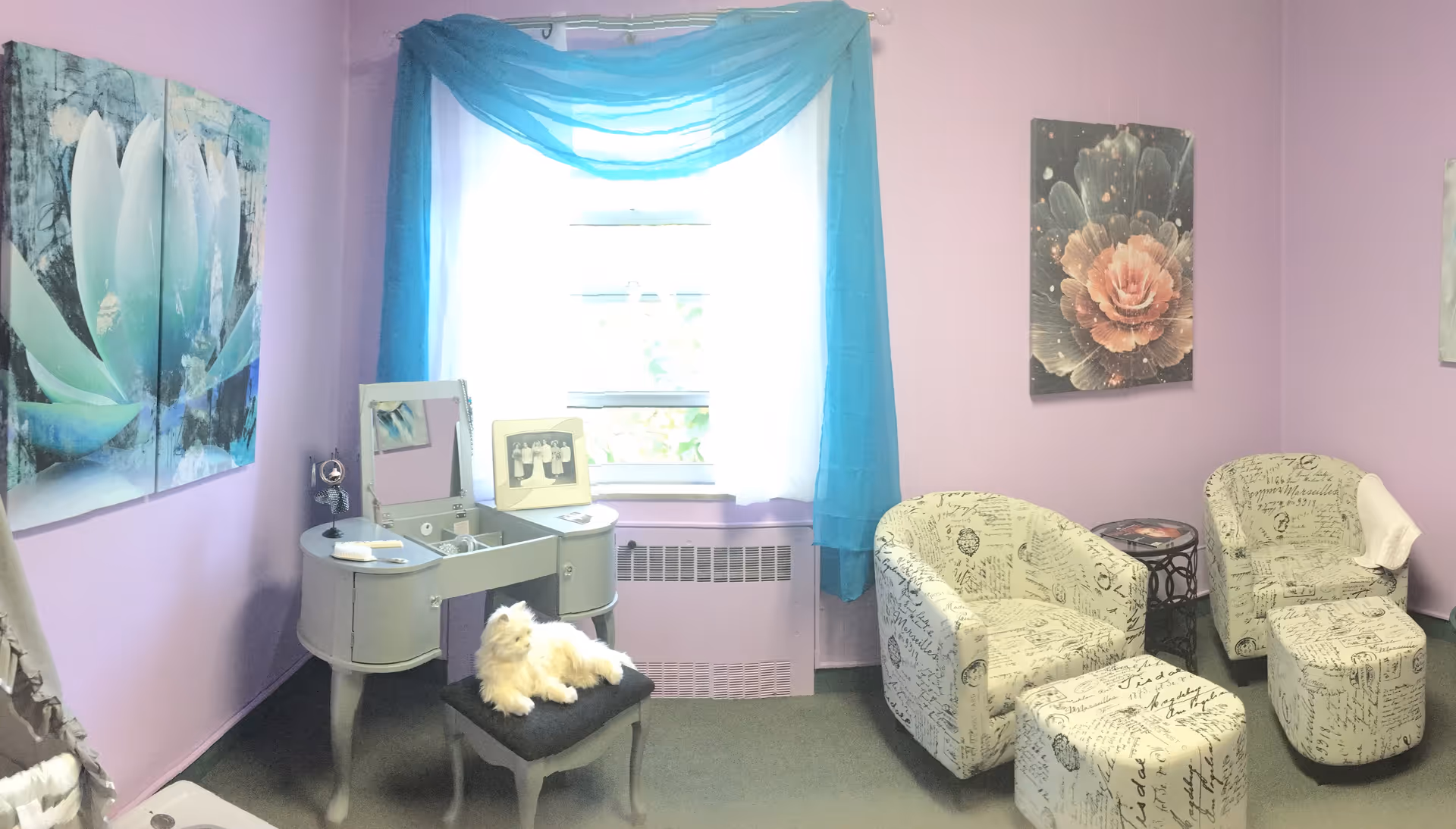A cozy room with lavender walls featuring a vanity table with a mirror and a small black cushioned stool with a white fluffy cat sitting on it. The window is adorned with sheer white curtains and a blue swag valance. Two armchairs and matching ottomans with script-patterned upholstery are arranged near a small round side table. Floral artwork hangs on the walls.