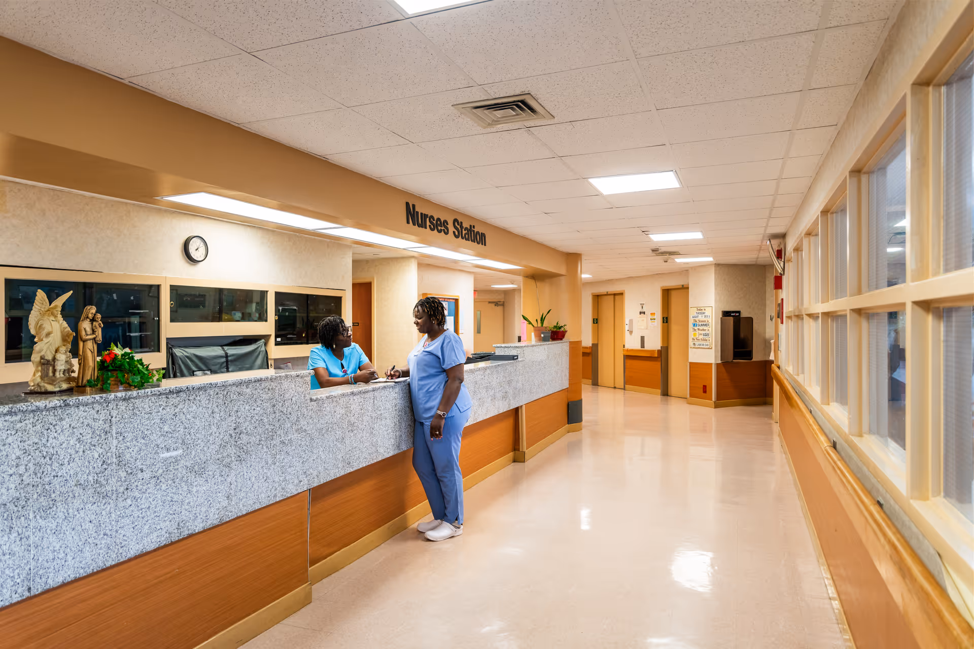 A bright hospital or care facility corridor with a nurses station on the left. Two healthcare workers in blue scrubs are engaged in conversation at the nurses station counter. The corridor has beige walls, a polished floor, and windows on the right side. There are statues and plants on the counter, and a clock on the wall behind the nurses station.