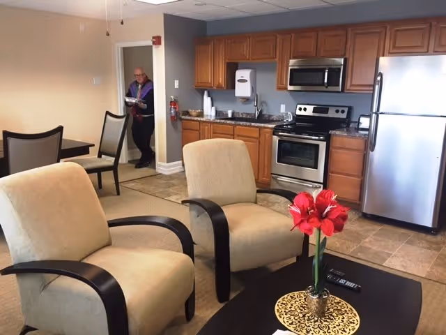 Interior view of a senior living facility's common area with a kitchen in the background featuring wooden cabinets, a stainless steel refrigerator, stove, and microwave. In the foreground, there are two beige armchairs with dark wooden armrests and a coffee table with a red flower arrangement. A man is standing in the doorway holding a book or folder.