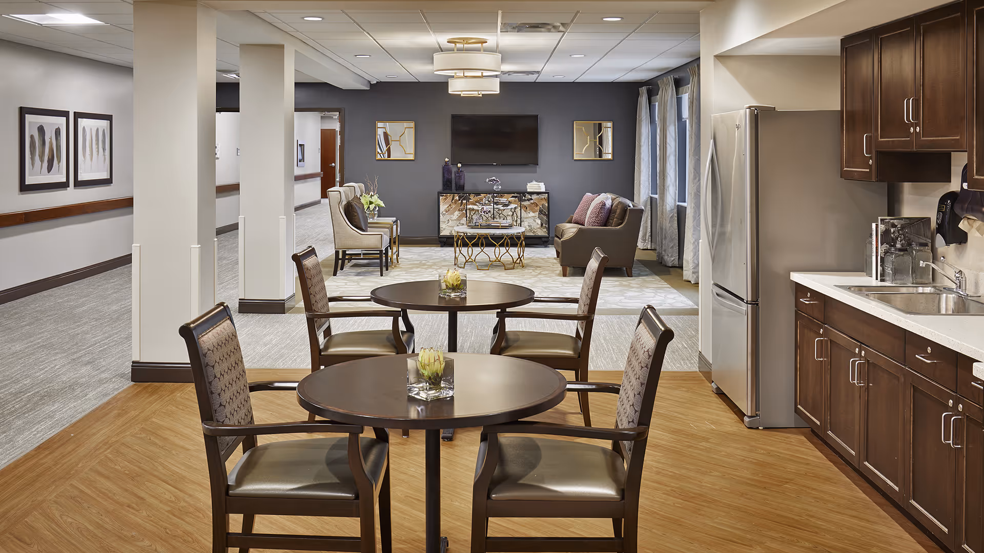 Open communal dining and lounge area with round tables and chairs, a kitchenette with stainless refrigerator on the right, and a seating area with a TV in the background.
