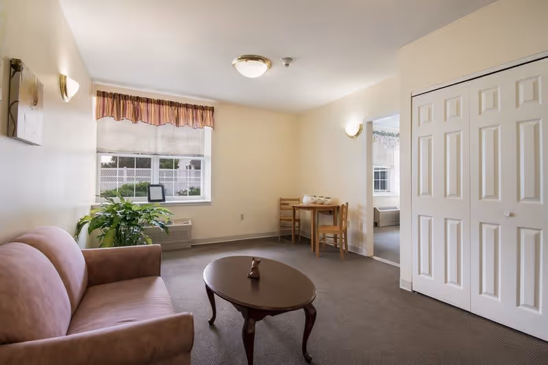 A small living room with a pink upholstered sofa on the left, a dark wooden oval coffee table in the center with a small decorative item on it, a green potted plant near a window with blinds and a striped valance. In the background, there is a small wooden dining table with two chairs and a tea set on top. The walls are light-colored, and there is a white double-door closet on the right side. The room is carpeted and well-lit with ceiling and wall lights.