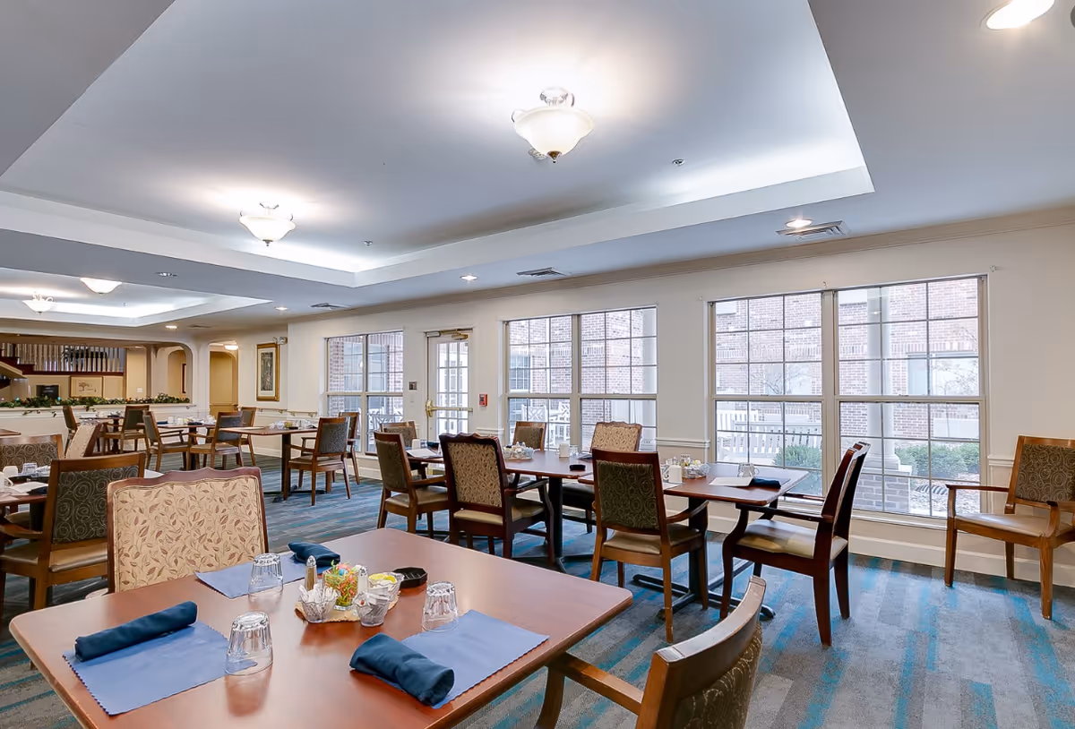 A bright and spacious dining room in a senior living facility with multiple wooden tables and chairs. Each table is set with blue placemats, folded blue napkins, glasses, and small condiment containers. Large windows along one wall allow natural light to fill the room, and the ceiling features recessed lighting and decorative light fixtures.
