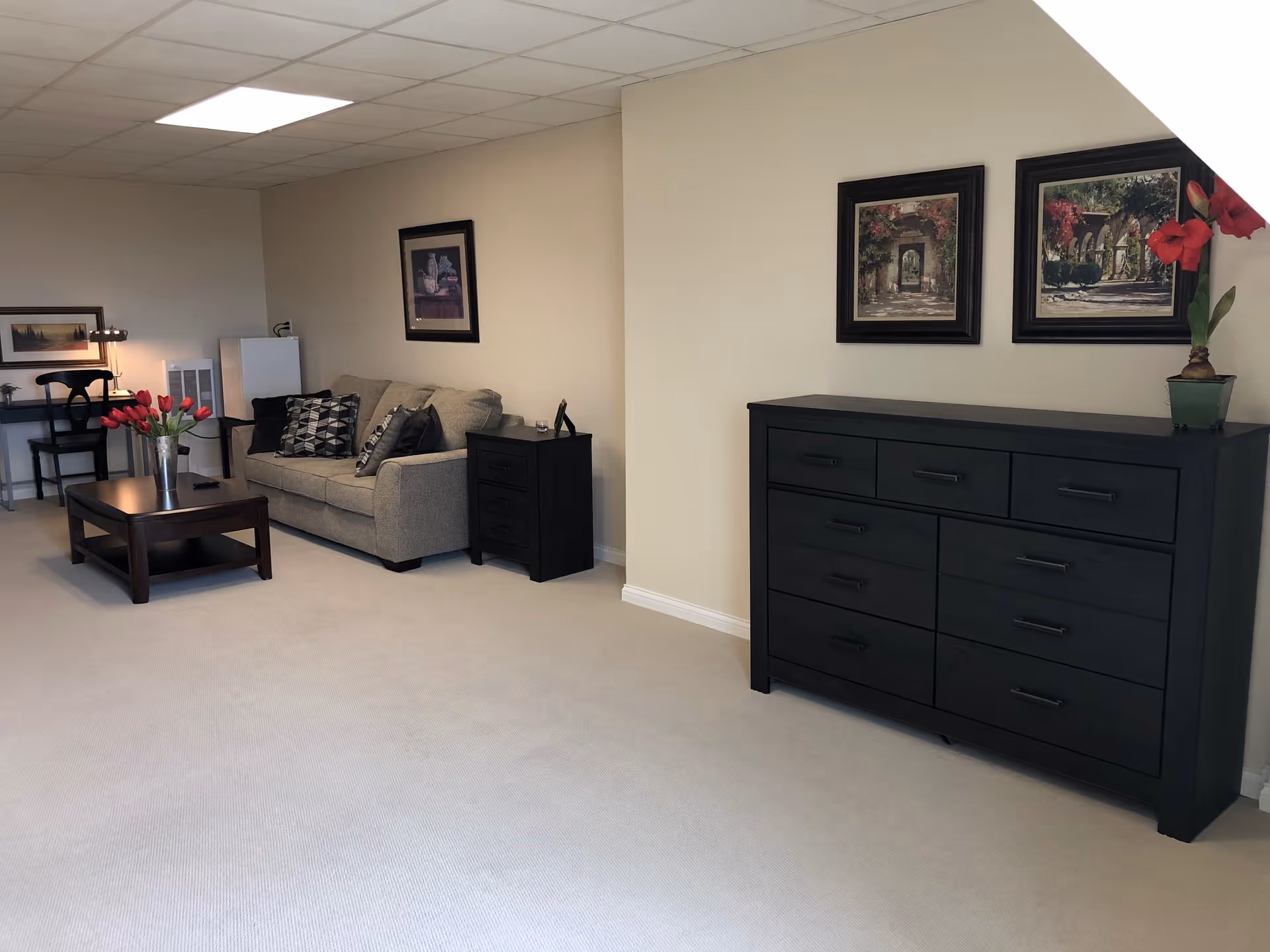 Bright furnished living room with a sofa, coffee table, dark dresser, framed artwork, and a small desk.