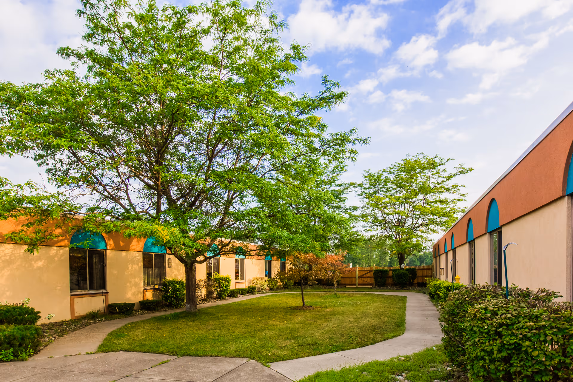 Sunny courtyard with green trees and a curved walkway between two single-story buildings with teal awnings.
