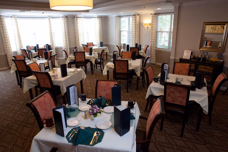 Dining room with multiple tables set with white tablecloths, place settings, menus, and chairs under soft lighting.