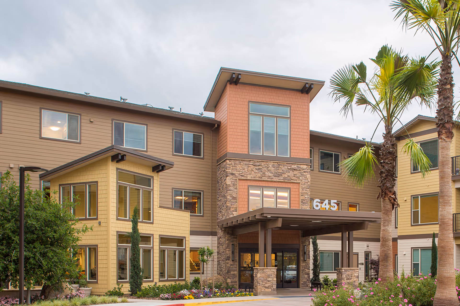 Exterior view of Prairie City Landing senior living facility showing a three-story building with a covered entrance, large windows, stone and wood siding, palm trees, and landscaped greenery.