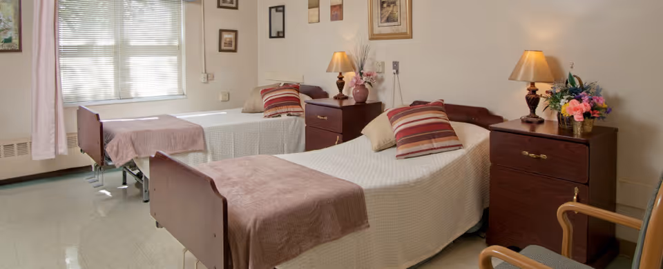 A bright room with two single beds, each with white bedspreads, brown blankets at the foot, and striped pillows. Between the beds are two wooden nightstands with lamps, a vase with flowers, and framed pictures on the wall. A window with blinds lets in natural light.